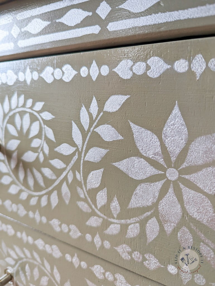 Close-up of the Faux Bone Inlay Stencil Hand-Painted Vintage Chest of Drawers, showcasing an intricate white floral and geometric pattern on a pale olive green background. The design features leaves, flowers, and circular motifs, giving a detailed, decorative appearance to the surface.