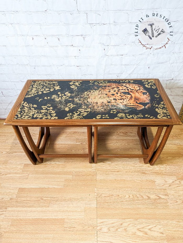 A G Plan Astro Long John Teak Nest of Tables featuring rectangular wooden tables with curved legs and an artistic top surface design. The detailed design showcases a leopard amid a pattern of gold-colored leaves on a dark background, all set against a light wooden floor.