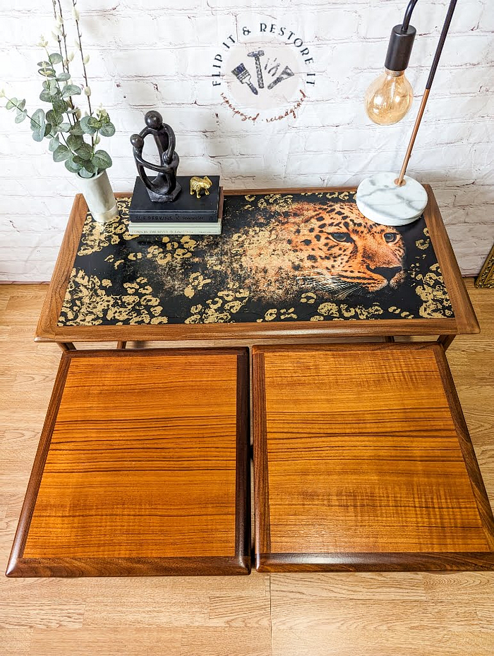 A G Plan Astro Long John Teak Nest of Tables, intricately painted with a leopard design on its surface, holds a black sculpture, a book, a small figurine, and a modern lamp. Below the table are two wooden storage units. A plant is placed to the left of the tables on the floor.