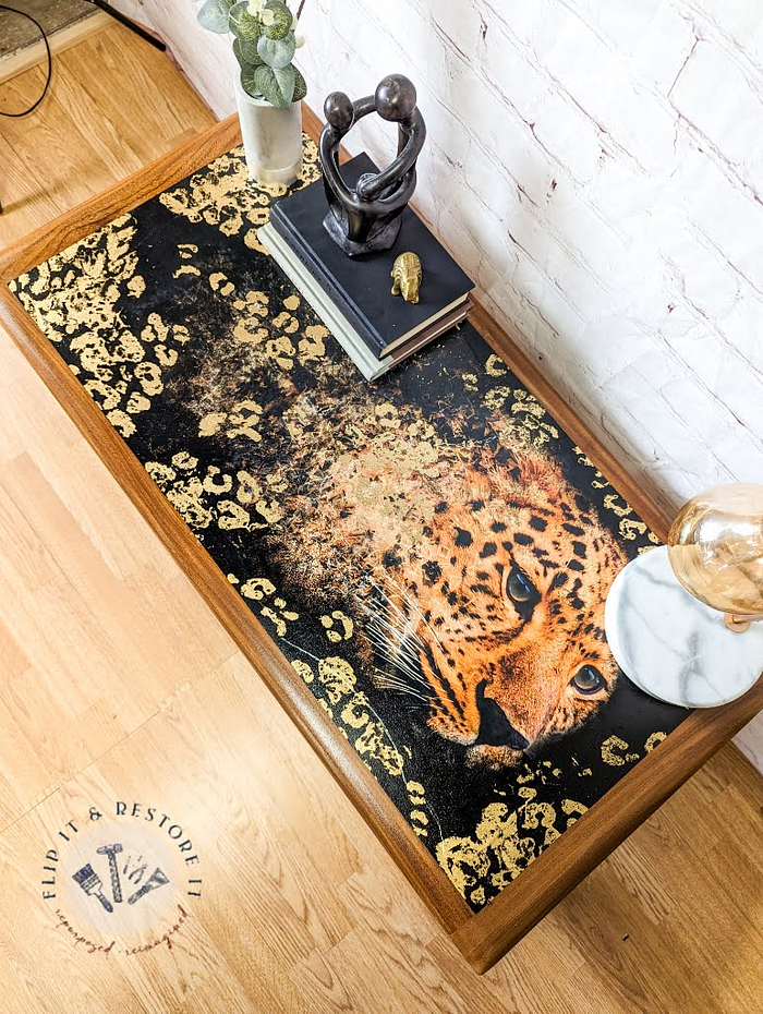 A G Plan Astro Long John teak coffee table featuring a detailed leopard image on its surface, surrounded by gold accents. On the table are books, a small figurine, a potted plant, and a marble-topped item. The floor is wooden, and the wall behind is white brick.