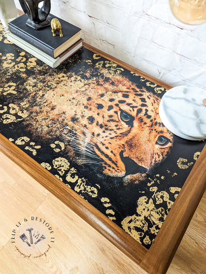 A set from the G Plan Astro Long John Teak Nest Of Tables with a wooden surface showcasing a striking design of a leopard's face merging into an artistic golden pattern. The table edges have a light wood finish, and it's adorned with books, decorative items, and a small round white marble piece.