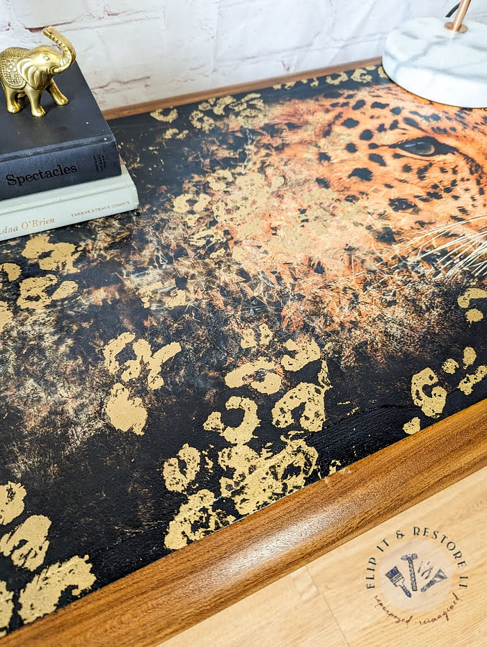 The G Plan Astro Long John Teak Nest Of Tables boasts a wooden table top adorned with a striking black and gold leopard print design. On the table, you'll find a selection of books alongside a small gold elephant figurine. The scene is set against the backdrop of a white brick wall, with the "Flip It & Restore It" logo prominently displayed in the bottom right corner.