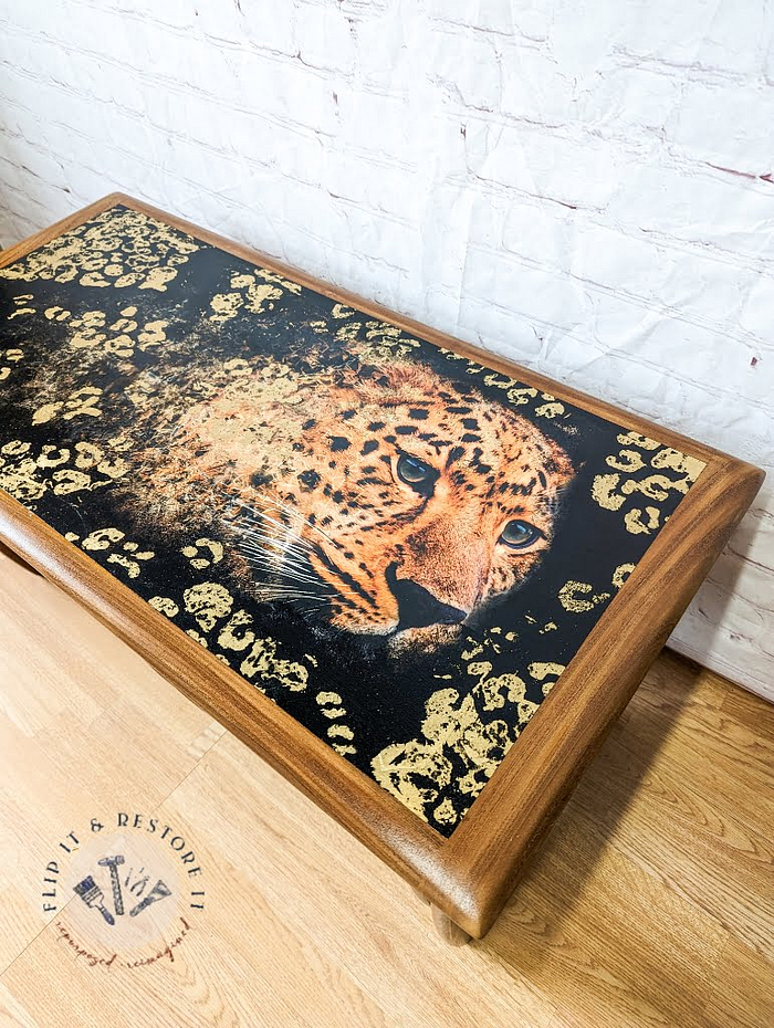 A G Plan Astro Long John Teak Nest of Table with a stunning painted design on the surface, showcasing a leopard's face encircled by an intricate pattern of spots. The table is set on a light-colored floor against a white brick wall, with the "Flip it & Restore it" logo visible in the bottom left corner of the image.