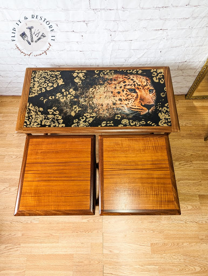 Vintage wooden side table from the G Plan Astro Long John Teak Nest Of Tables collection, featuring intricate artwork of a leopard on its black tabletop. Below the tabletop is a single drawer front. The scene includes wooden floors and a light-colored brick wall in the background, with part of a golden framed mirror visible on the right side.