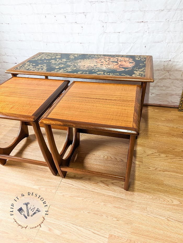 The G Plan Astro Long John Teak Nest Of Tables, a set of three wooden nesting tables, is stacked together on a wooden floor against a white brick wall. The largest table features a black tabletop adorned with a floral pattern. A logo in the bottom left corner reads "FLIP IT & RESTORE IT.