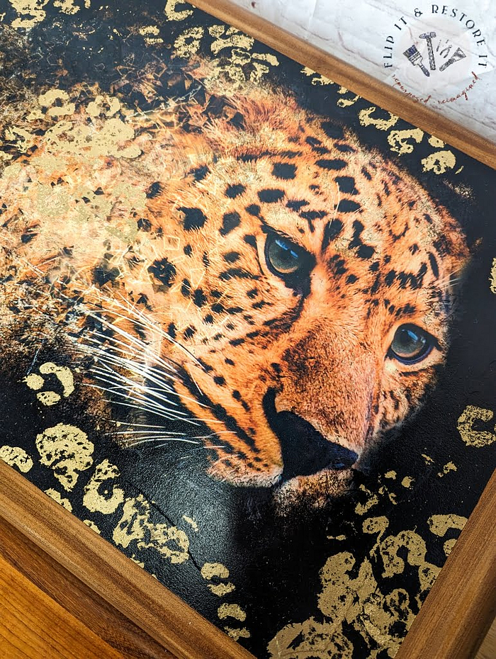 The G Plan Astro Long John Teak Nest of Tables features an intricate, close-up depiction of a leopard's face, accentuated by its intense blue eyes. This striking artwork combines realism with abstract elements and is set against a textured backdrop with golden highlights to enhance the leopard's mesmerizing features.