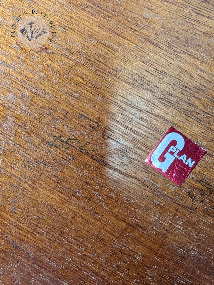 A section of a teak surface exhibits signs of fading, scratches, and wear. On the right side, a red and white "G Plan" sticker is visible. In the upper left corner, there is a circular "Build & Destroy Wares" logo. This piece is part of the G Plan Astro Long John Teak Nest Of Tables collection.