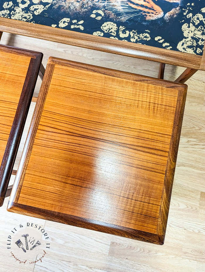 A close-up of the G Plan Astro Long John Teak Nest Of Tables alongside a matching wooden stool. The table features a sleek black surface adorned with a decorative edge pattern. The floor is light-colored hardwood, and in the bottom left corner, there is a logo with the tagline "Flip It & Restore It".