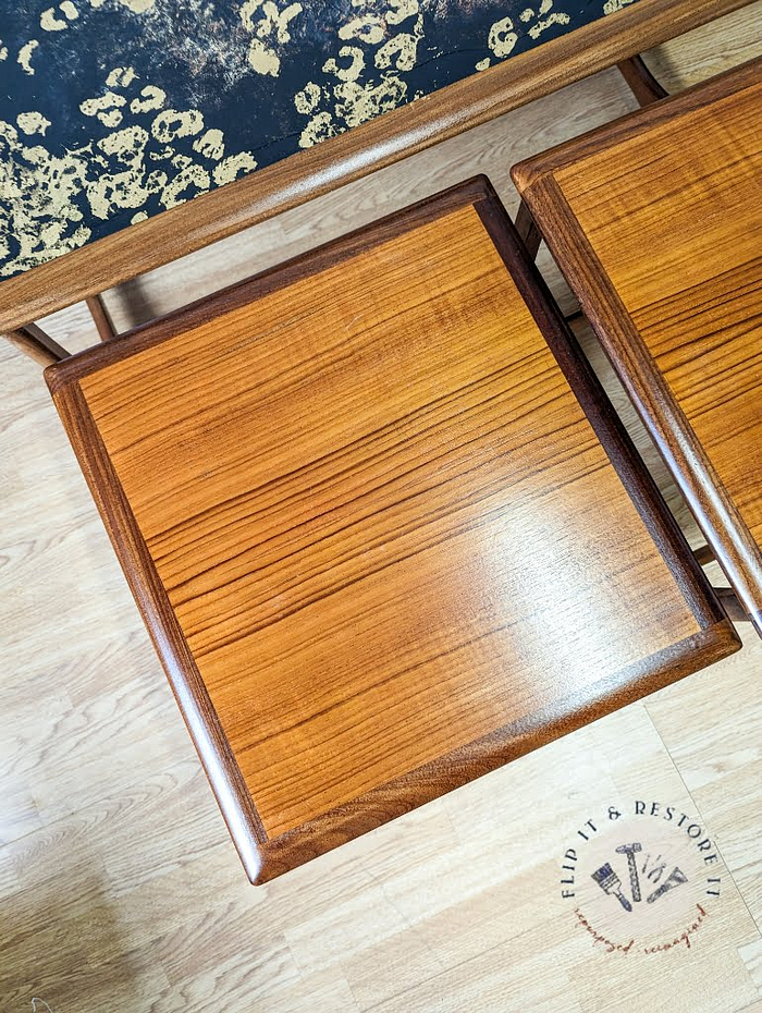 A close-up of the G Plan Astro Long John Teak Nest Of Tables reveals a polished wooden surface with a natural grain pattern. The table's finish gleams under the light, highlighting its intricate craftsmanship. In the background, part of another similar table from the same set and the edge of a dark, abstract-patterned object are also visible.