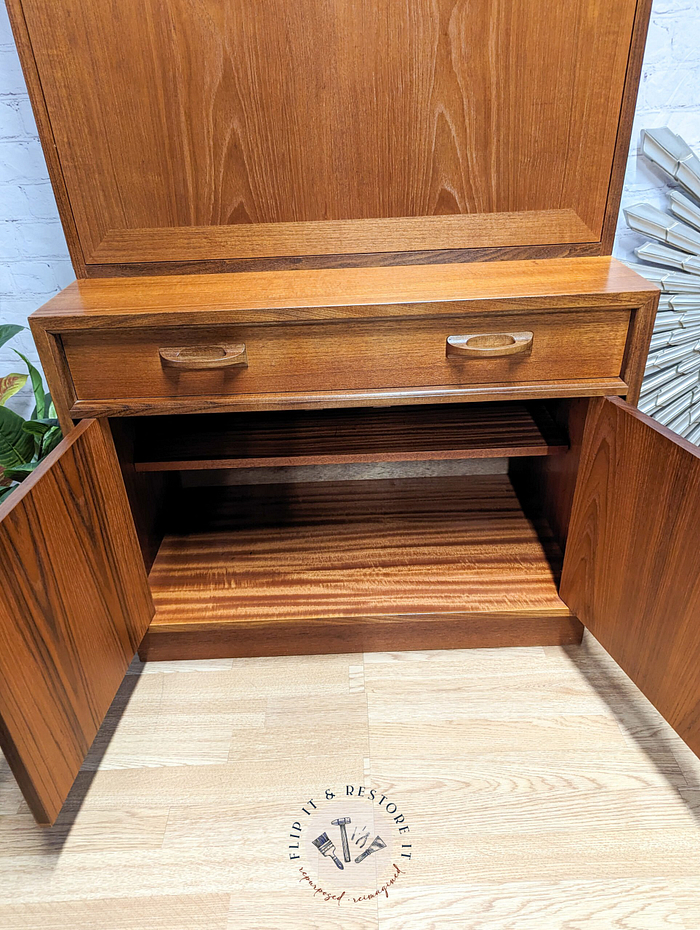 The G-Plan Sierra Tall Teak Bookcase/Drinks Cabinet Mid Century features a single drawer on top and double doors below, which open to reveal an interior shelf. The cabinet boasts a natural wood finish and is situated on a light wooden floor with a logo that reads "Flip it & Restore it.