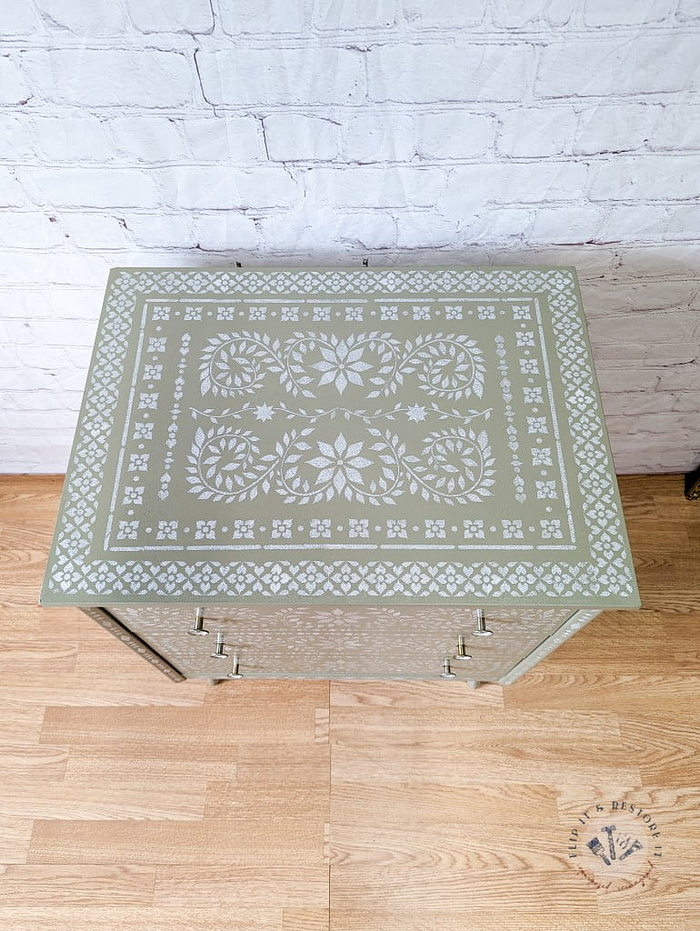 An overhead view of the Faux Bone Inlay Stencil Hand-Painted Vintage Chest of Drawers reveals its stunning green surface adorned with intricate white floral and geometric patterns. The chest stands on a light wooden floor, set against a white brick wall backdrop.