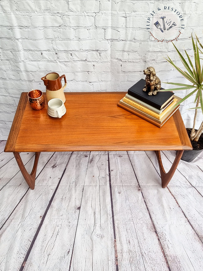 A G Plan Astro Mid Century Modern Vintage Teak Coffee Table from the 1960's with curved legs is placed against a white brick wall. On the table are books, a dog figurine, a potted plant, a white mug, a copper container, and a white pitcher with a wooden handle. The floor is wooden.