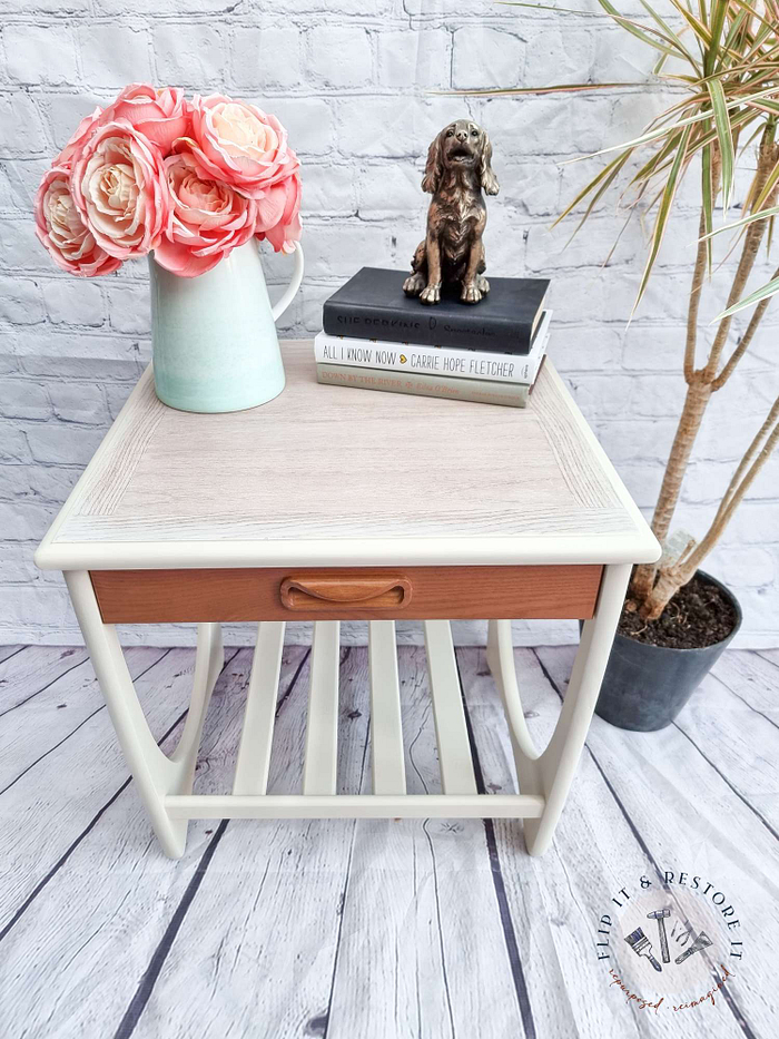 A Mid Century Beautility Drinks Cabinet from the 1960s, made of walnut and designed for use as a vintage cocktail bar, features a vase containing pink and white flowers, two stacked books, and a dog statuette. The cabinet sits on a wooden floor with a brick wall background and is accompanied by a potted plant on the right.