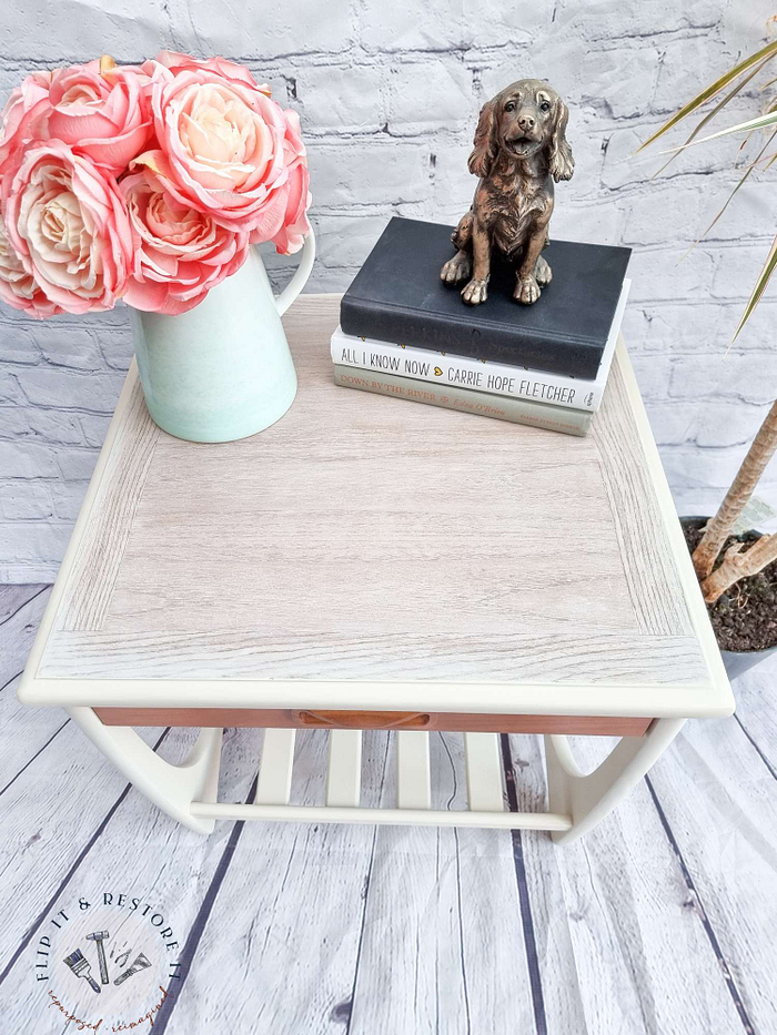 A small, cream-colored side table with a light wooden top, adorned with a white jug of pink roses, a stack of books, and a bronze dog figurine, stands beside the Mid Century Beautility Drinks Cabinet. The scene is set against a light wood floor and a brick wall background, with a leafy plant partially visible on the right.