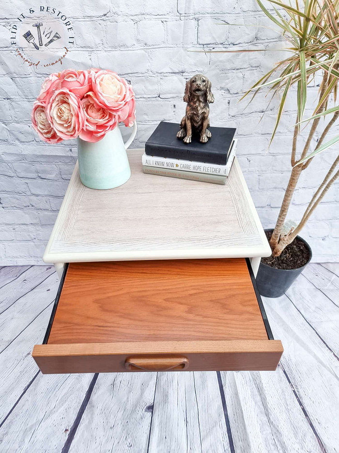 A compact Mid Century Beautility Drinks Cabinet from the 1960s, featuring a two-tiered design with elegant walnut surfaces. The top tier displays a vase filled with pink roses, a dog sculpture, and two books. The lower tier includes an open wooden drawer. A tall potted plant stands on the floor next to the cabinet, set against a white brick wall backdrop.