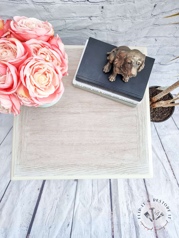 A Mid Century Beautility Drinks Cabinet from the 1960s, with a walnut finish and vintage charm, features a vase of pink roses and two stacked books, topped with a bronze dog figurine. This stylish cabinet rests on a wooden floor next to a tall plant, showcasing its light wood top adorned with a subtle decorative border.