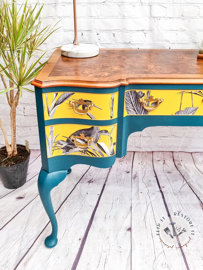 The Queen Anne Painted Burr Walnut Dressing Table/Vanity/Desk/Sideboard boasts vibrantly painted teal legs and yellow drawers, adorned with intricate black and white bird and foliage illustrations. Its top is a natural wood finish. A potted plant sits to the left on a white, wooden floor against a white brick wall background.