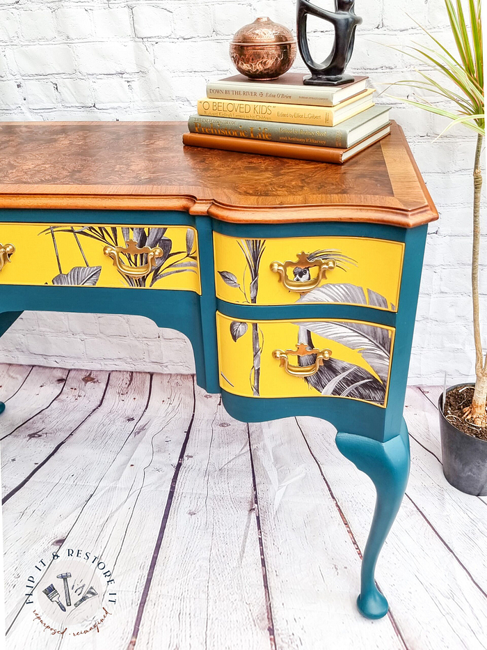 A vibrantly painted Queen Anne Painted Burr Walnut Dressing Table/Vanity/Desk/Sideboard featuring teal and yellow drawers adorned with floral and bird designs. The surface holds a stack of books, a decorative plant, and a small sculpture. The scene is set against a white brick wall and wooden floor.