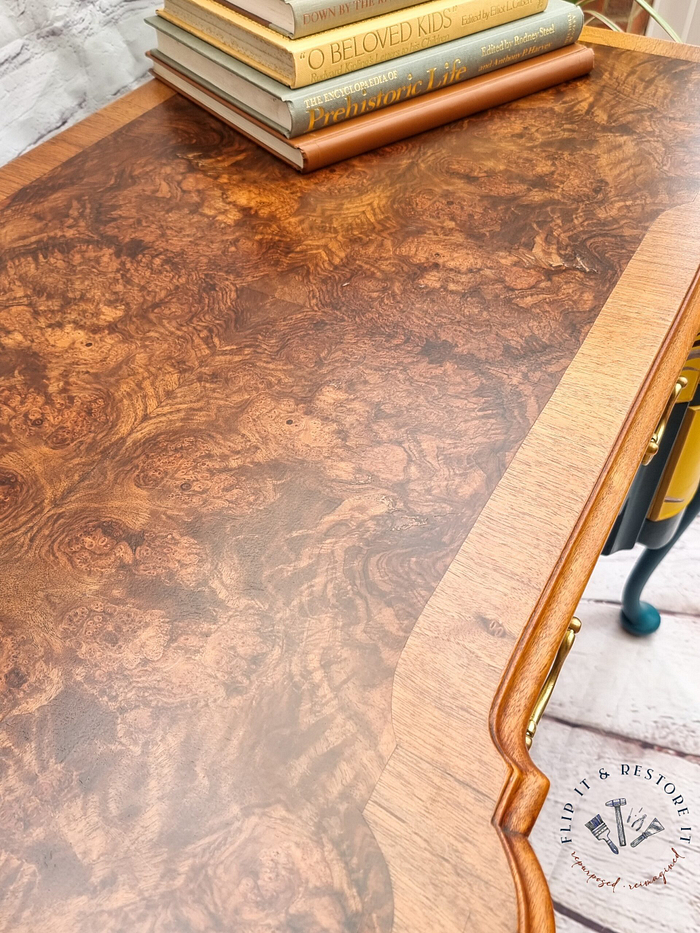 A Queen Anne Painted Burr Walnut dressing table, vanity, desk, or sideboard with a richly textured, polished surface. Books are neatly stacked on one side, including titles like "Prehistoric Life" and "Loved Kids." The edge of the desk features intricate details, and a logo in the corner reads "Flip It & Restore It.