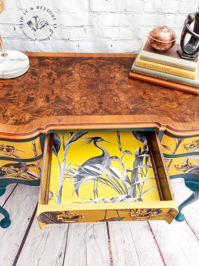 A Queen Anne Painted Burr Walnut Dressing Table/Vanity/Desk/Sideboard against a white brick wall. The desk features intricate grain patterns and a drawer that is open, revealing a vibrant yellow interior with a black and white bird and reed design. Books, including one titled "Beloved Kids," are stacked on top.