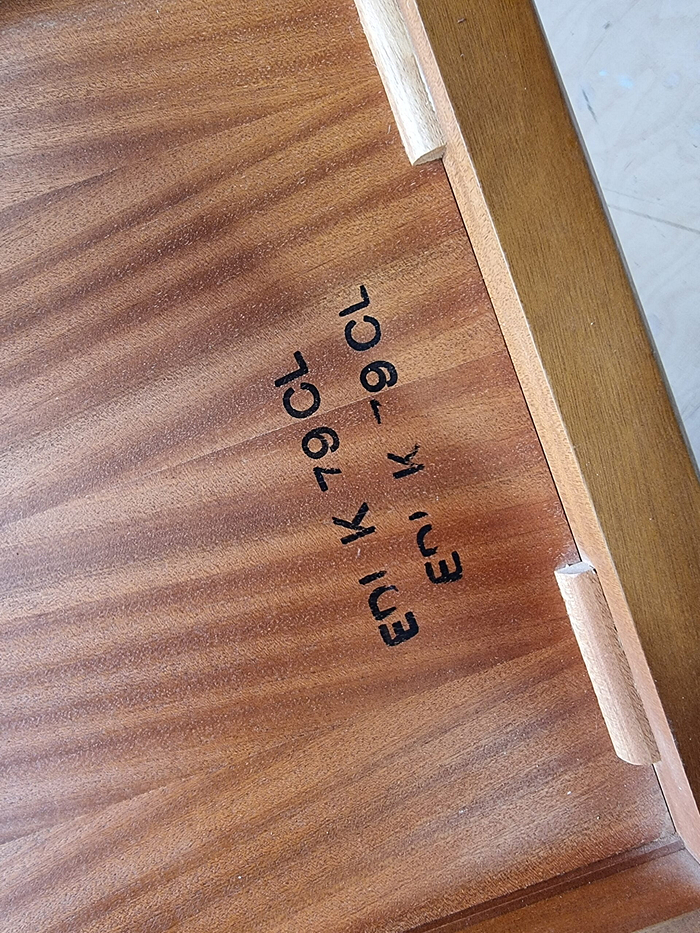 A close-up of the interior of a wooden drawer from the Mid Century Teak Nest of Tables - Coffee Table and Two Side Tables, showcasing its unique wood grain pattern. The text "EN I K-79 CL" is stamped in black ink on the inner surface. The drawer features wooden guides and is partially opened, revealing its construction details.