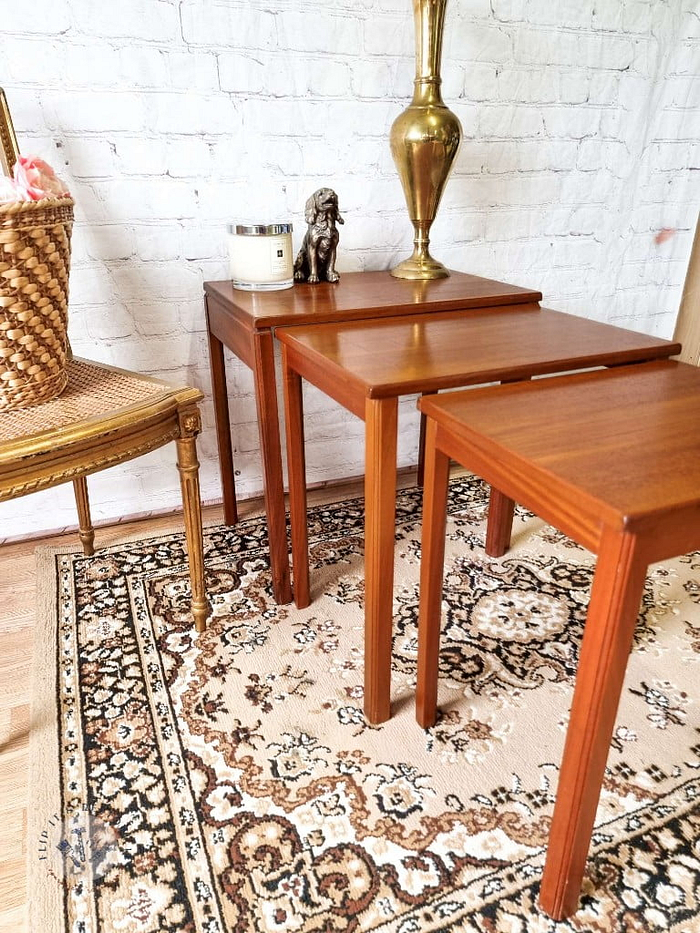 The Ercol Saville Ash Long Coffee Table with Smoked Glass Mid Century Vintage is arranged on a patterned rug. A brass lamp, a white candle, and a small bronze figurine are displayed on the table. An ornate wicker basket is placed on a decorative chair to the left. The background features a white brick wall.