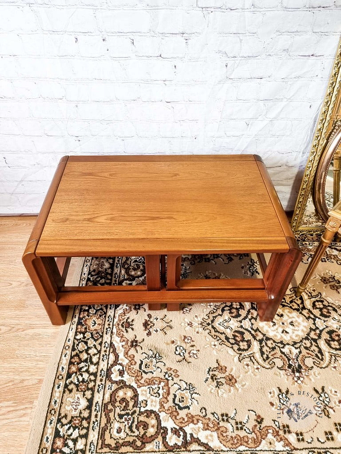 A Mid Century Teak Nest of Tables, featuring a rectangular coffee table and two side tables with open sides, sits on a patterned rug. In the background, there's a white brick wall and a gold-framed mirror leaning against it. The floor is light wood, and part of a chair with a cushion is visible to the right.