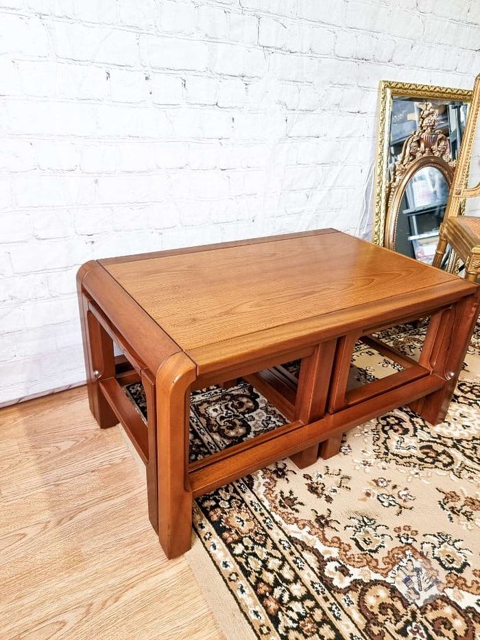 A Mid Century Teak Nest of Tables, featuring a coffee table and two side tables, sits on a patterned beige rug. Next to it, leaning against a white brick wall, are ornate gold-framed mirrors and a chair with similar wood design.