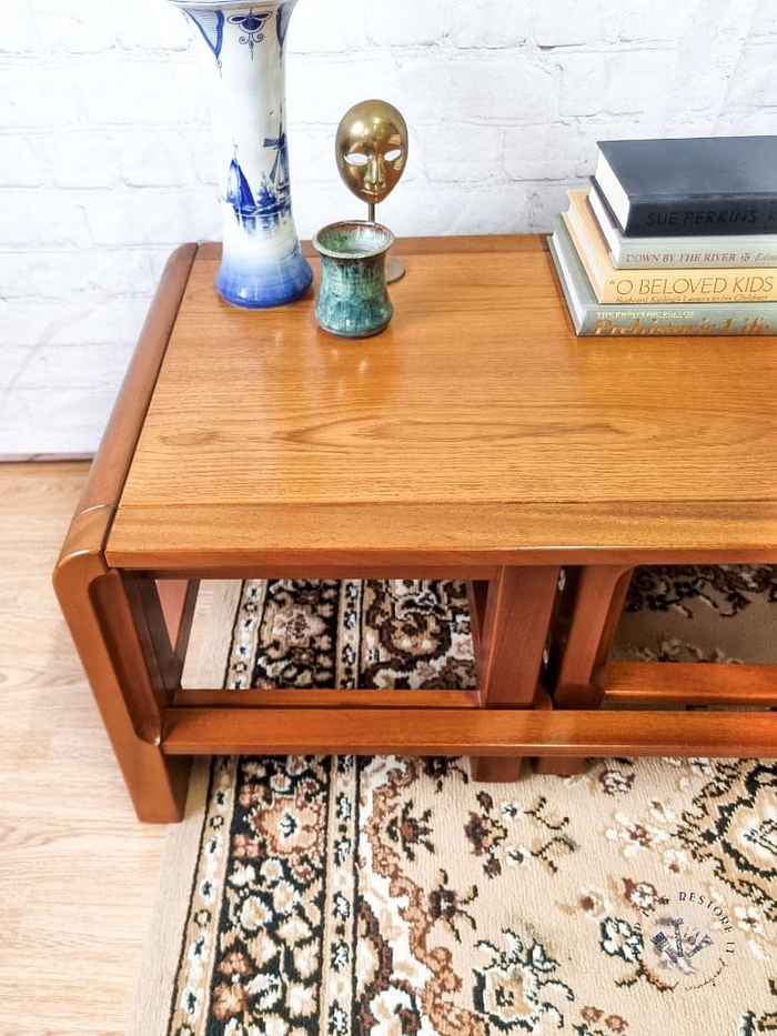 The Mid Century Teak Nest of Tables - Coffee Table and Two Side Tables display a striking blue and white vase, a petite decorative sculpture, a green candle holder, and several stacked books. The tables rest on a beige patterned rug set against a white brick wall background.