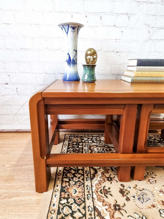 A Mid Century Teak Nest of Tables - Coffee Table and Two Side Tables set with a glossy finish is placed against a white brick wall. On the tables are books, a blue and white porcelain vase, a small green statue, and another small gold sculpture. A patterned rug is on the floor under the tables.