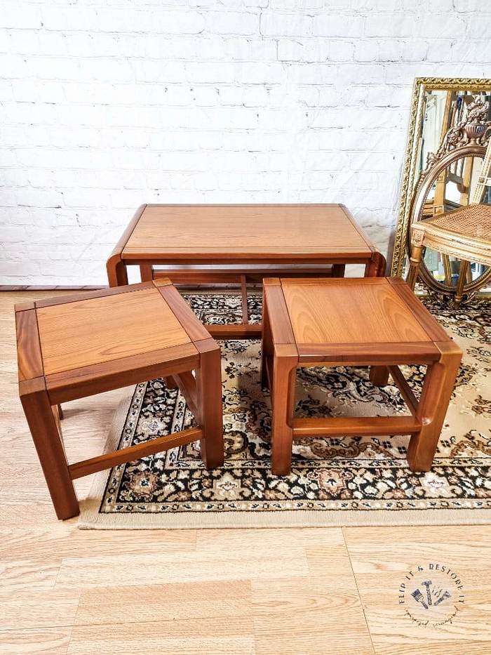 A set of three Mid Century Teak Nest of Tables, featuring a clean and minimalist design, are placed on a patterned area rug. The two smaller square side tables are nested under the larger rectangular coffee table. A gilded ornate chair and a large decorative mirror adorn the background against a white brick wall.