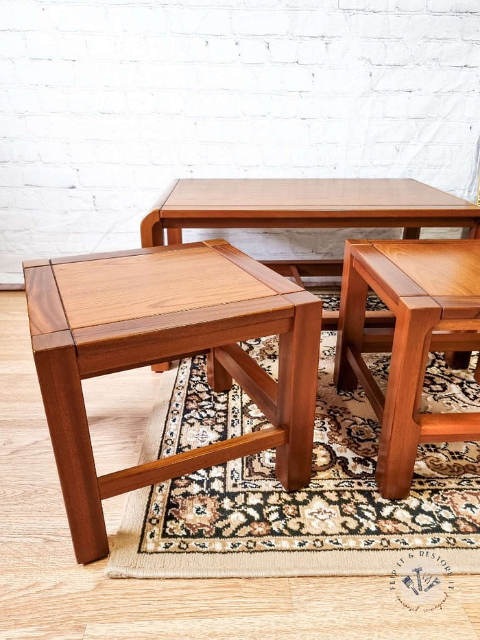 This image showcases the Mid Century Teak Nest of Tables, featuring a coffee table and two side tables with clean lines and a minimalist design. The two side tables are positioned in the foreground on an ornate patterned rug, while the coffee table is in the background against a white brick wall. The floor is wooden.