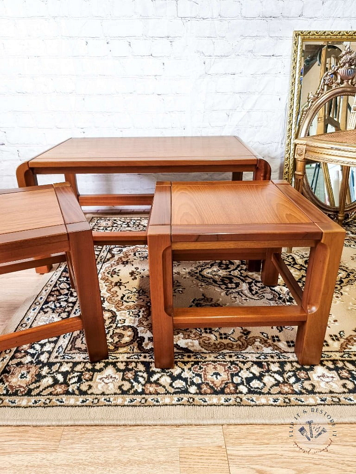 A Mid Century Teak Nest of Tables - Coffee Table and Two Side Tables are arranged on a patterned rug in front of a white brick wall. Two rectangular side tables flank the larger rectangular coffee table. A gold-framed mirror and a small decorative piece are visible in the background.