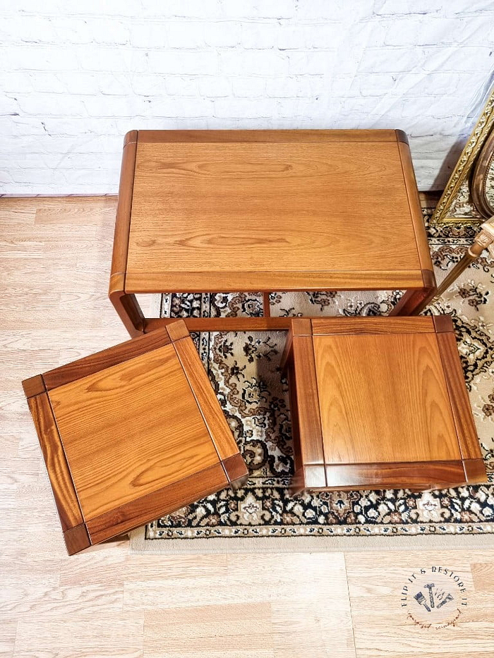 The Mid Century Teak Nest of Tables, consisting of one coffee table and two side tables, are displayed on a light wood floor against a white brick wall. Arranged on top of a small, decorative patterned rug, the rectangular coffee table is complemented by the two square side tables. A part of a framed mirror can be seen to the right.