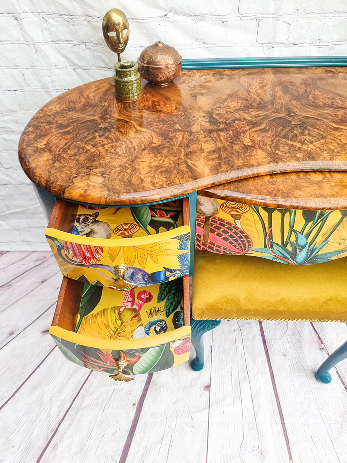 The Queen Anne Painted Burr Walnut Bedroom Set features a vintage-style dressing table/vanity with a polished curved surface. The open drawers are painted with vibrant floral and animal designs. The set includes yellow upholstered seating and is adorned with small decorative items, including a golden mask and a small copper container.