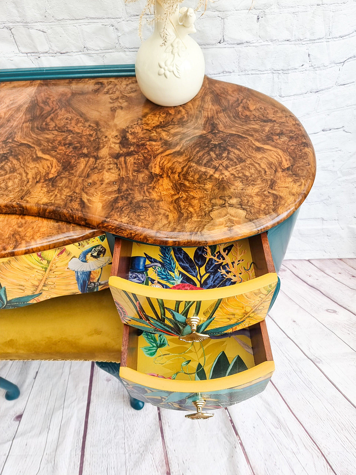 A beautiful dressing table from the Queen Anne Painted Burr Walnut Bedroom Set, featuring a wooden frame with a marbled top and vibrant, nature-themed linings inside the partially open small drawers. The interior showcases yellow hues adorned with leafy and bird designs. A cream-colored vase decorates the top of this elegant piece, set against a white brick backdrop.