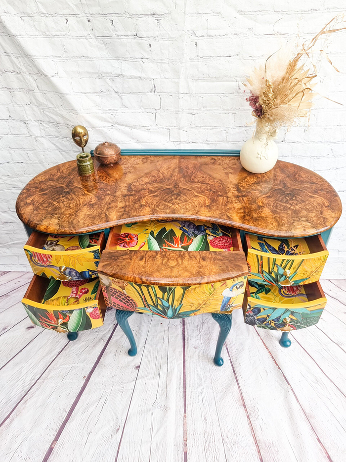 A Queen Anne Painted Burr Walnut Dressing Table from the Bedroom Set, featuring six open, floral-patterned drawers. The top is adorned with two small sculptures and a vase with dried flowers. The legs of the table are painted blue-green. It is set against a backdrop of a white brick wall and light wood flooring.