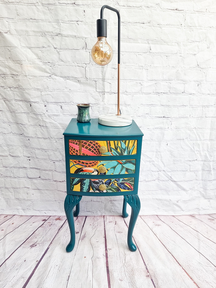 A small, teal-colored wooden dressing table from the Queen Anne Painted Burr Walnut Bedroom Set, featuring intricate tropical-themed artwork on its drawers, stands on elegantly carved legs. A decorative pottery piece and a modern, minimalist lamp with a round bulb sit on top. The backdrop is a white brick wall and a light wooden floor.