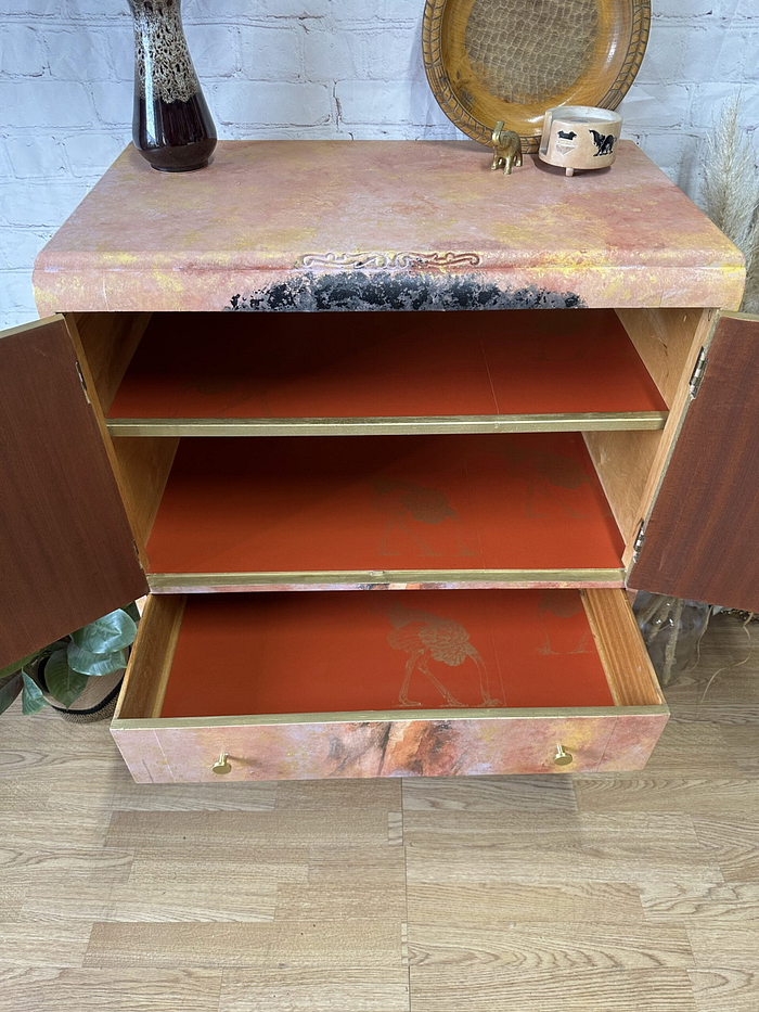 A hand-painted cabinet, standing tall as a versatile linen cupboard, has its doors open to reveal three striking red-painted shelves. On top of the upcycled cupboard sits a charming decorative vase filled with dried flowers, accompanied by a round tray, a small container adorned with an animal design, and a petite elephant figurine. The cabinet stands gracefully on a wooden floor.