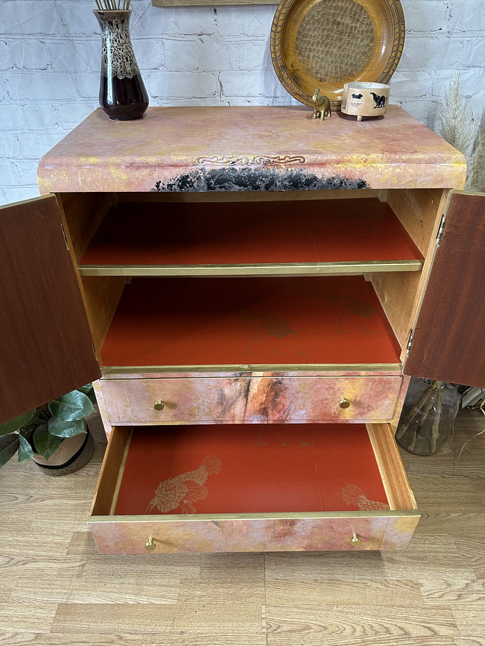 A tallboy hand-painted cabinet that features an upper section with an open shelf and a lower section with drawers, all painted red inside. The exterior showcases a textured pink design with black marks. It is surrounded by decorative items including a tray, vase, and small plants. Suitable as linen cupboard or linen press, this upcycled piece can be used as bedroom storage, kitchen storage, or clothes storage.