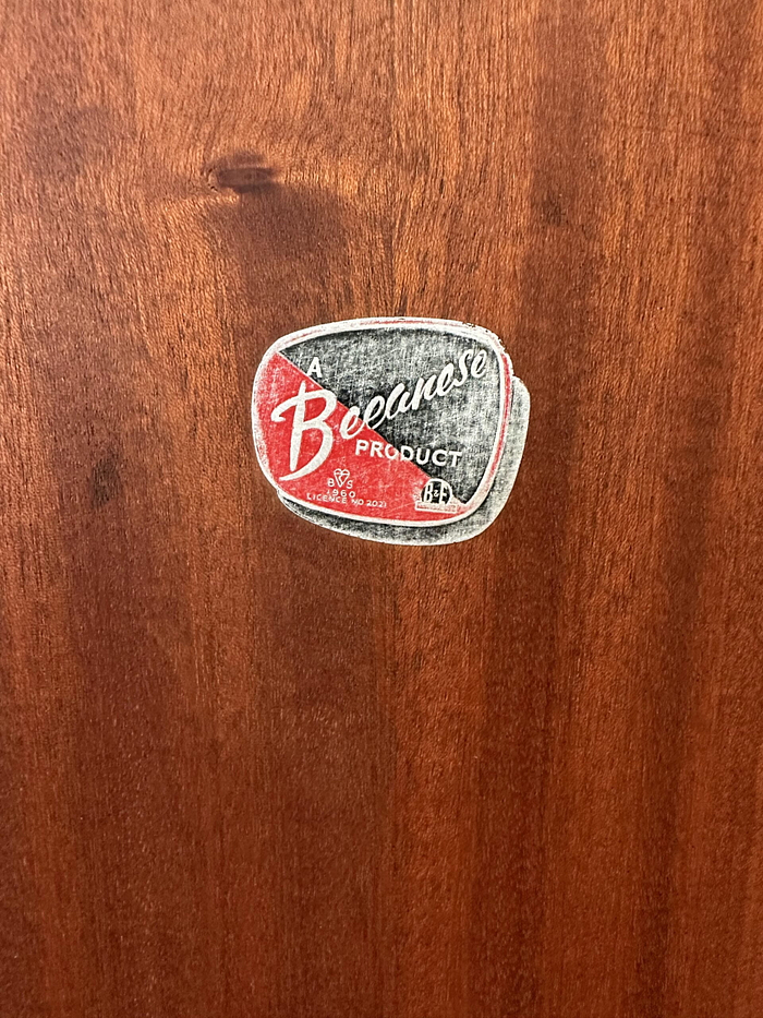 A wooden surface featuring an oval-shaped label in the center that has a red and white color scheme and displays the text "A Beanese Product" along with some other smaller, unclear text. The label exhibits a vintage, retro design. This piece is part of the Hand-painted cabinet, Tallboy, Linen Cupboard, Linen Press, Upcycled Cupboard collection and can be used for Bedroom Storage, Kitchen Storage, or Clothes Storage.