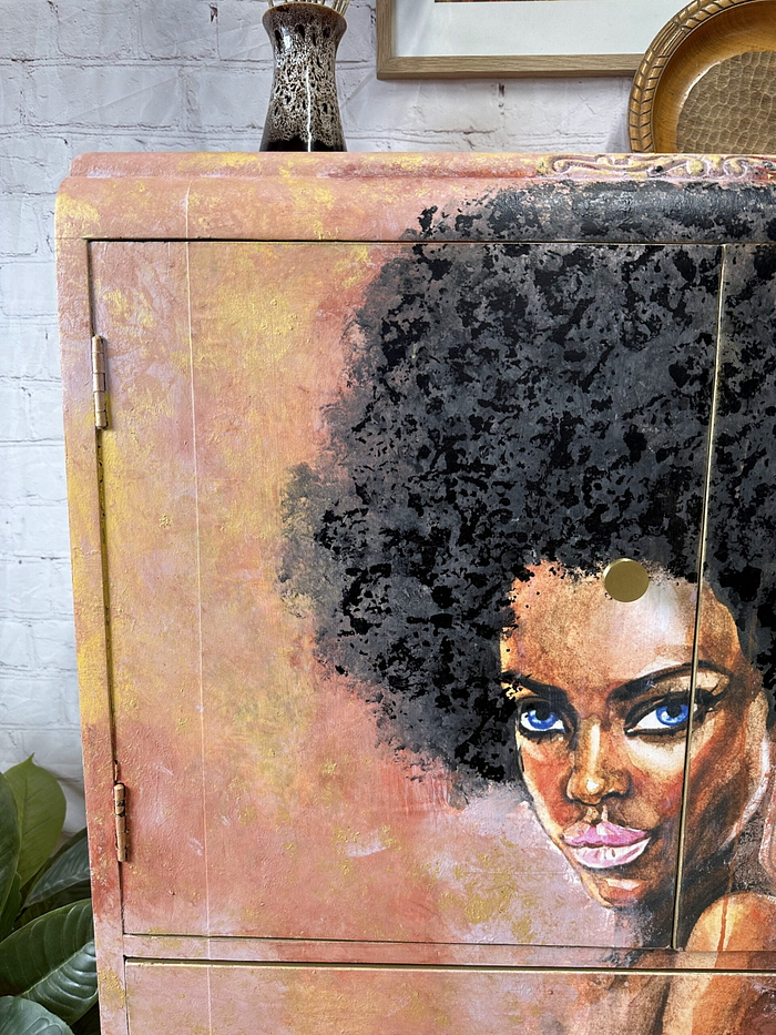 A Tallboy hand-painted cabinet featuring a portrait of a woman with a large afro hairstyle. The artwork adorning the door of this upcycled linen cupboard beautifully illustrates her eyes, nose, and lips in vibrant detail. A small ceramic vase graces the top of the cabinet, and a plant is visible to the left. This versatile piece can be used as bedroom storage, kitchen storage, or clothes storage.