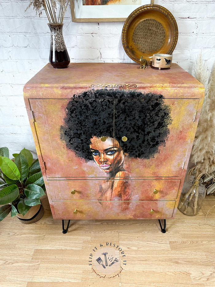 A Hand-painted Cabinet adorned with artwork of a woman with an afro on its doors, featuring a light brown finish, gold handles, and metal legs. Surrounding it are houseplants and decorative items like a small vase and a wooden bowl.