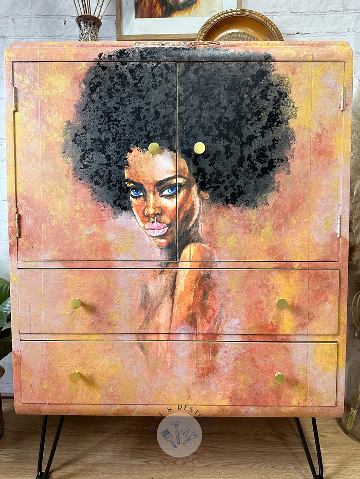 The Hand-painted Cabinet, featuring a vibrant portrait of a woman with an afro hairstyle, is adorned with warm hues of orange, yellow, and gold. This upcycled Tallboy Linen Cupboard boasts four drawers and two doors with round handles. The background showcases a wooden floor and some decor.