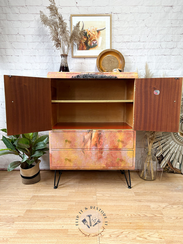 A Hand-painted Tallboy cabinet stands on a wooden floor against a brick wall, with its doors open to reveal empty shelves. Decorative items, plants, and a framed artwork of a highland cow are arranged on and around the upcycled cupboard.