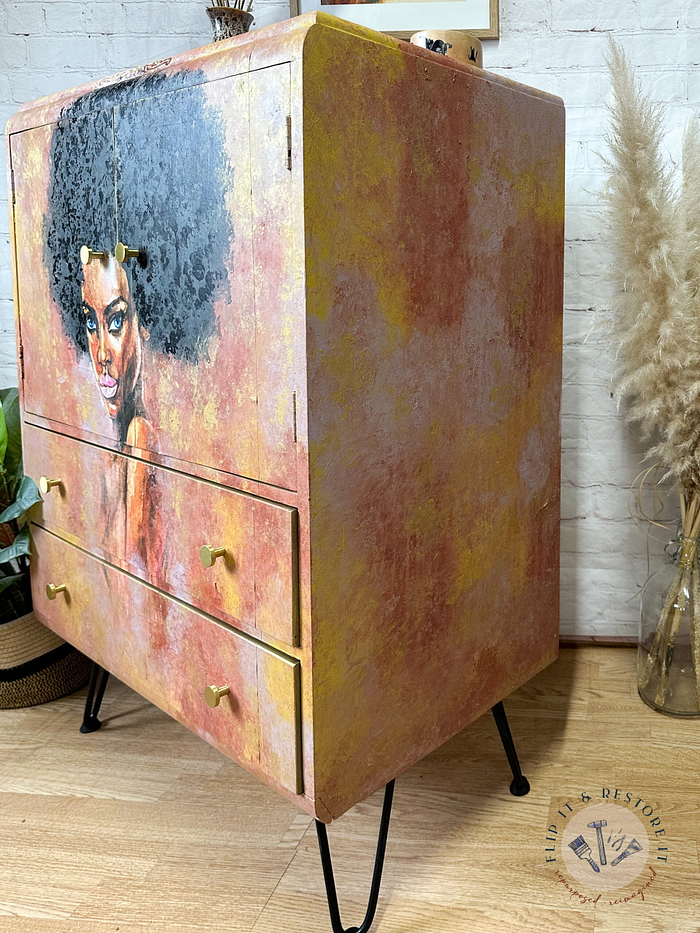 A hand-painted linen cupboard with two drawers is finished in warm tones and showcases an artistic portrait of a woman with curly black hair on the left side. The tallboy cupboard stands on sleek black metal legs and is set against a white brick wall, with dried grasses in a vase visible on the right.