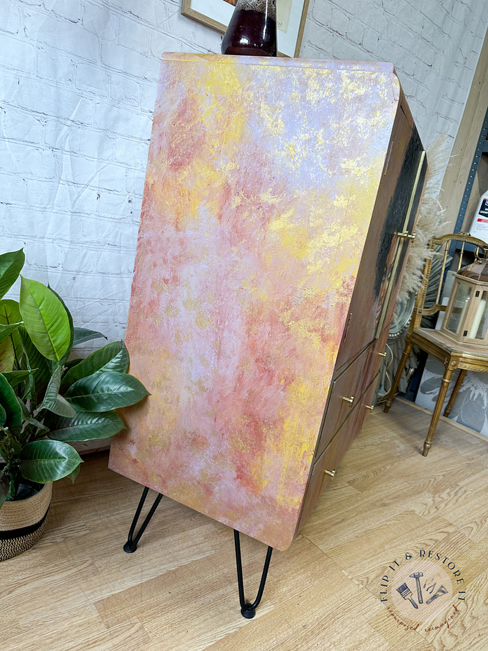 A side view of the Hand-painted cabinet, Tallboy, Linen Cupboard. It features abstract pastel colors of pink, yellow, and white. The upcycled cupboard stands on black hairpin legs with brass handles and is placed against a white brick wall. A green potted plant is situated to the left, with small decor items visible in the background.