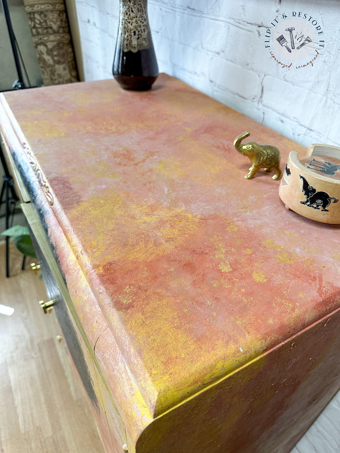 A close-up of a decorated Hand-painted cabinet with a distressed pink, orange, and yellow finish. On top are a ceramic vase with intricate patterns, a small gold elephant figurine, and a wooden box with a black design. A logo reading "Flip it & Restore it" is on the wall.