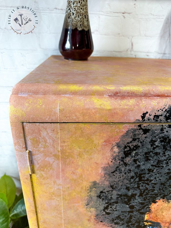 Close-up of a hand-painted Tallboy cabinet, featuring a textured finish in vibrant yellow and pink tones. A vase with intricate brown detailing sits on top. The background showcases a white brick wall. In the upper left corner, the "Flip It + Restore It" logo is partially visible.