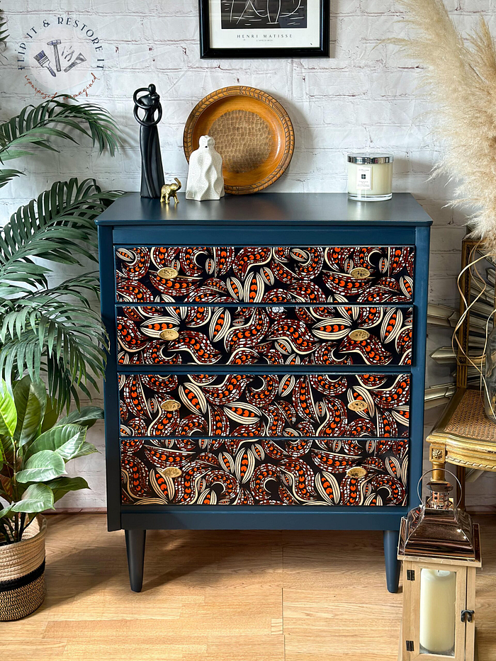 A dark blue chest of drawers with intricate red and orange paisley patterns sits against a white brick wall. It is adorned with decorative items, including a gold plate, a candle, and small sculptures. Surrounding the chest are potted plants and a lantern.